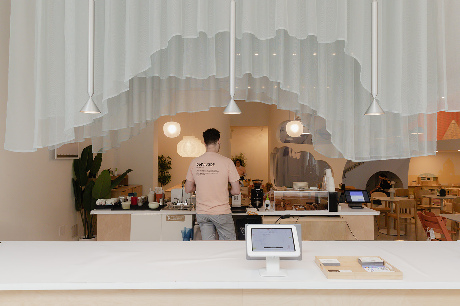 The Det'hygge café counter at House of Wonder with hanging pendant lights and a barista preparing coffee.